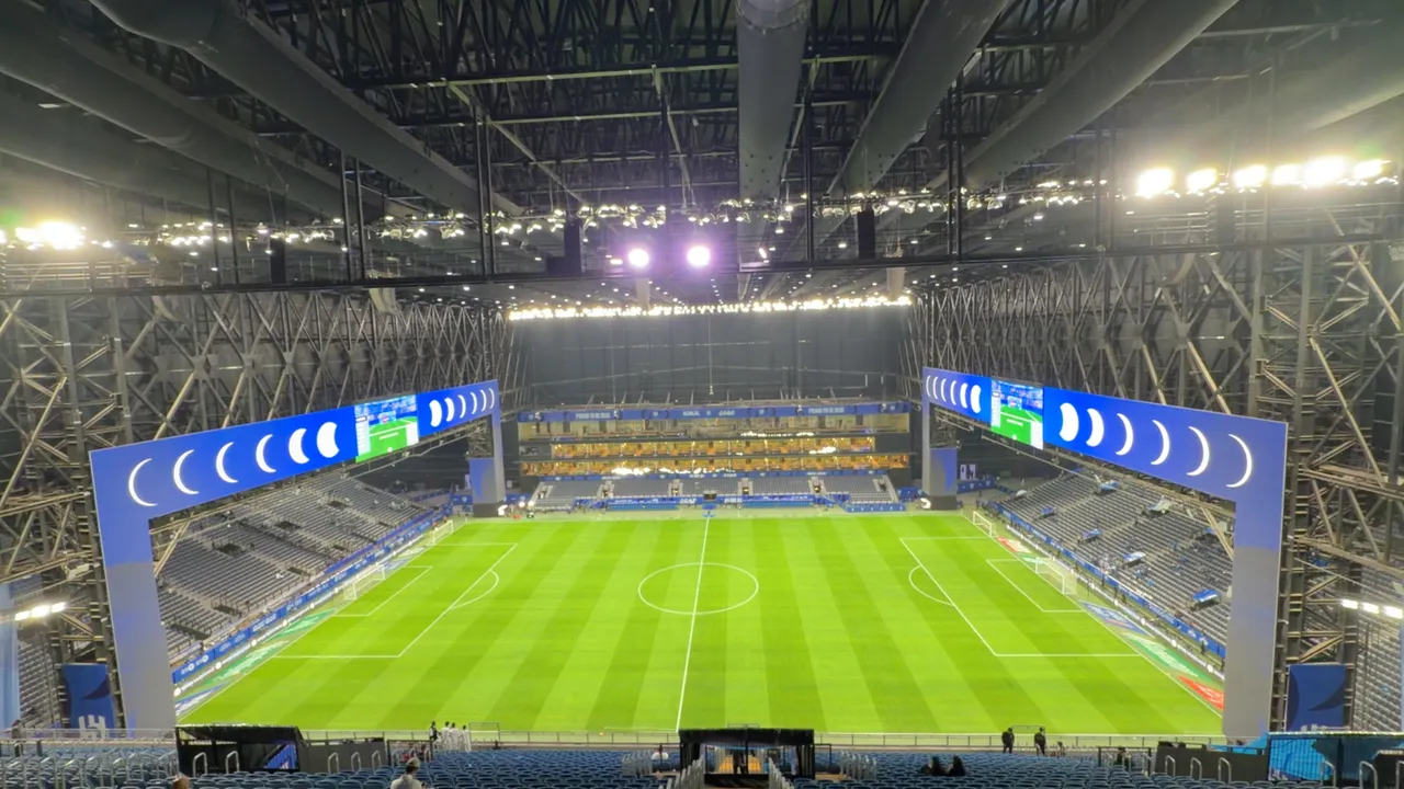The full pitch at Kingdom Arena from an elevated centre position, both LED arches lit blue and the steel roof trusses and lighting rig spanning the sealed bowl overhead
