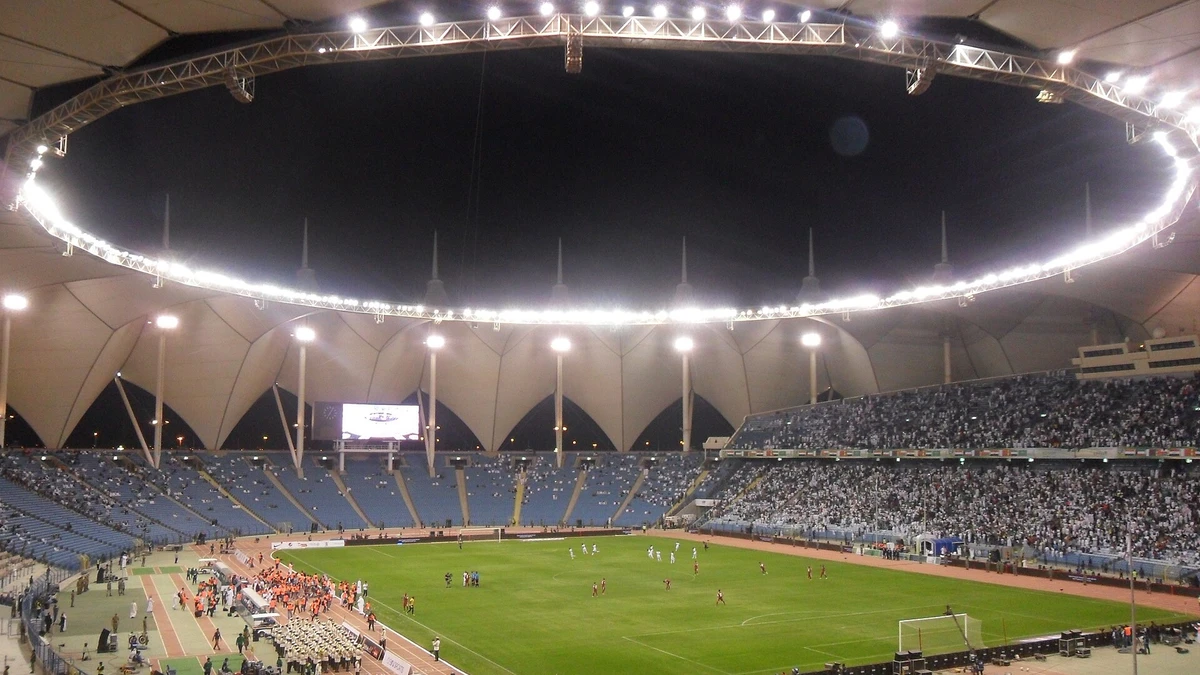 King Fahd Sports City Stadium in Riyadh at night, its tensile roof lit from below and the athletics track still ringing the pitch