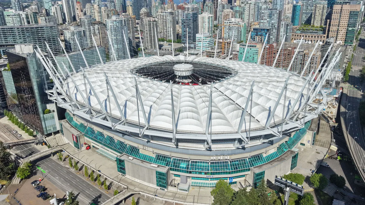 Aerial view of BC Place stadium in Vancouver with the white retractable roof closed, glass-towered downtown skyline rising directly behind the bowl