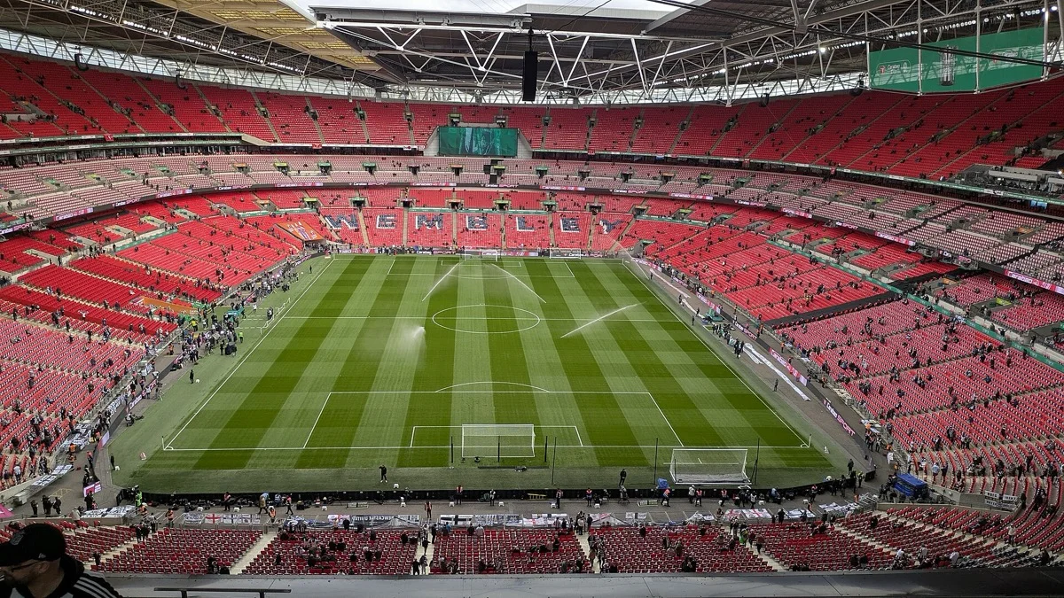 The bowl of Wembley Stadium seen from the upper tier before a match, the pitch being watered and the WEMBLEY lettering across the far stand