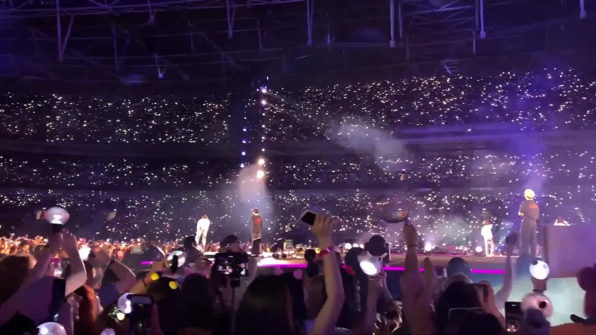 A packed stadium concert crowd at Wembley Stadium at night, a sea of phone lights facing the stage