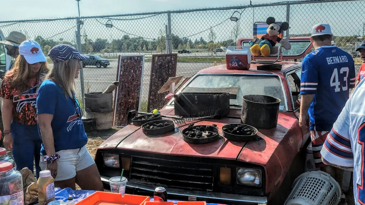 Buffalo Bills fans in jerseys tailgating around a red Ford Pinto with cast-iron pans grilling on the hood in the Orchard Park lots