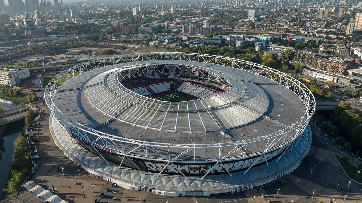 London Stadium from the air in its West Ham configuration, the claret and blue bowl inside the white lattice roof with the City of London skyline beyond