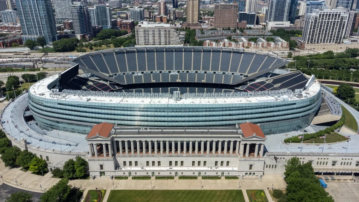 Aerial view of Soldier Field in Chicago, the 2003 glass and steel bowl rising behind and far above the low 1924 limestone colonnade, with the city skyline beyond