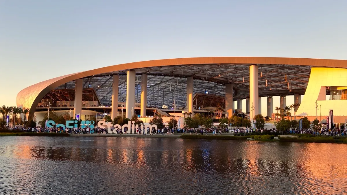 SoFi Stadium at sunset, its name spelled out in large freestanding letters on the plaza beside the lake