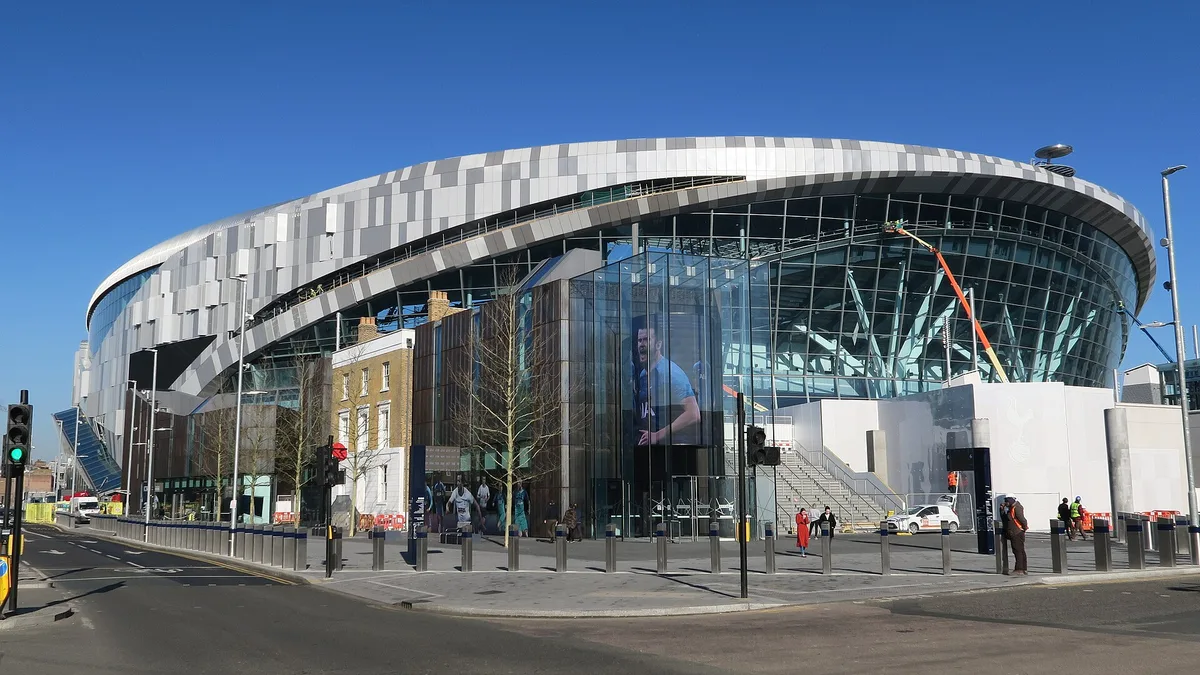 Tottenham Hotspur Stadium seen from the High Road, its glazed corner and curved metal facade rising above the street in north London