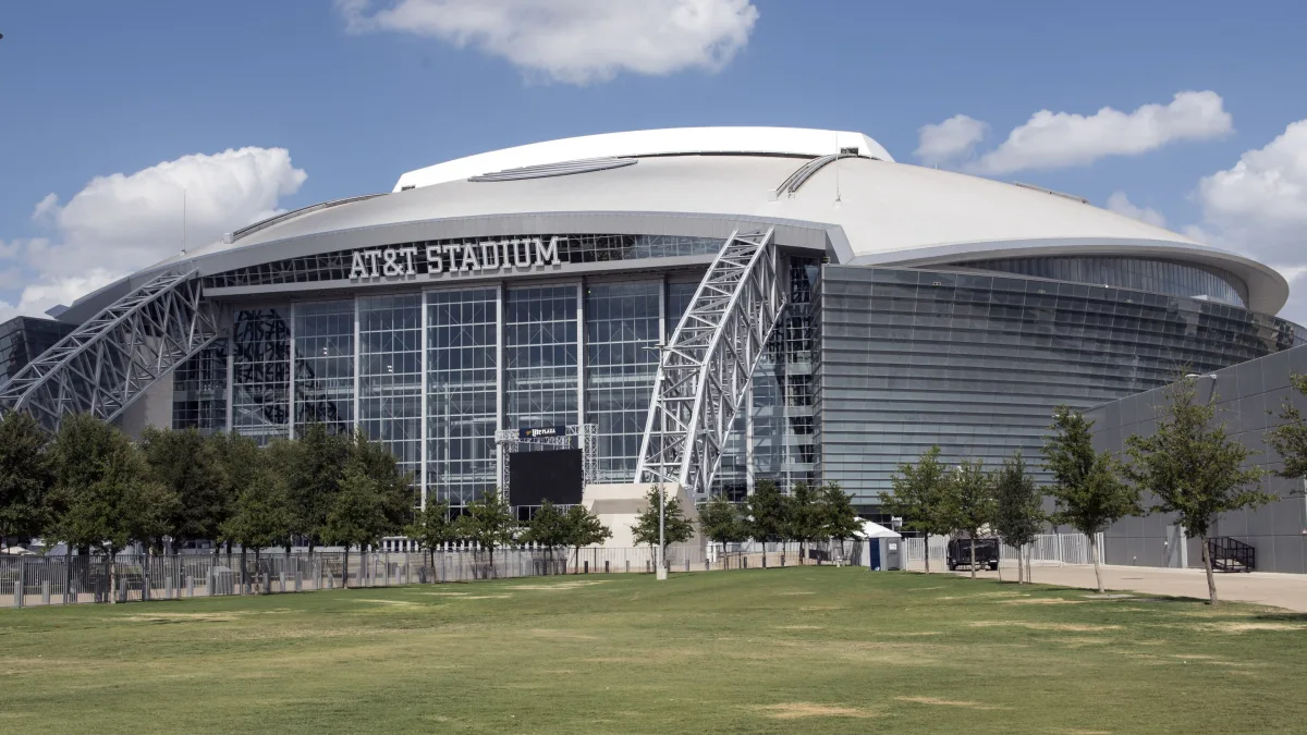 AT&T Stadium in Arlington, Texas, its arched steel trusses and name sign above the glass facade, seen across the lawn on a clear day