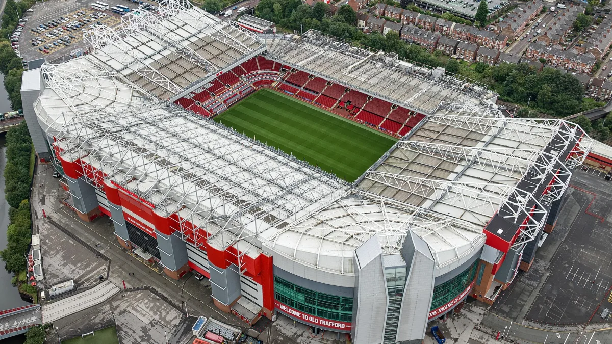 Aerial view of Old Trafford, the cantilever roofs over all four stands with the pitch and red seats at the centre, the East Stand frontage reading Welcome to Old Trafford and The Theatre of Dreams, and the Bridgewater Canal beyond